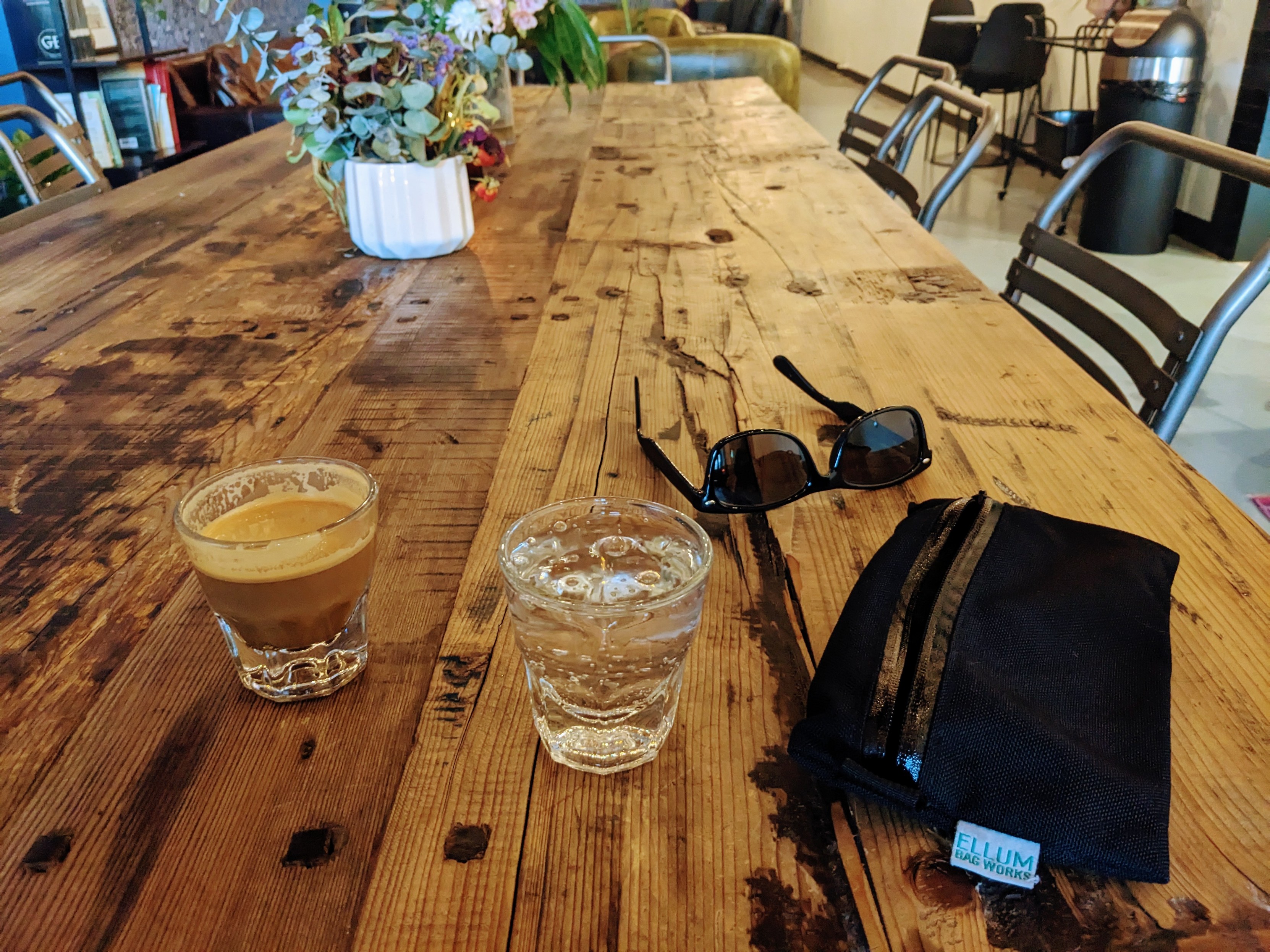 A long wooden table. On it rests a cortado, sparkling water, sunglasses, and a bike wallet/pouch made by Ellum Bag Works