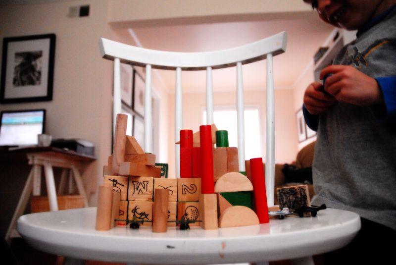 a white wooden chair with vertical bars on the backrest, which is being used as a makeshift play surface. On the chair’s seat, there is a carefully arranged structure made from wooden blocks of various shapes, colors, and sizes. Some of the blocks have letters, numbers, and symbols on them, while others are plain or painted in red and green. The structure resembles a small cityscape or castle.

In the background, a child wearing a gray and blue long-sleeve shirt is seen with a joyful expression, seemingly engaged with the block arrangement