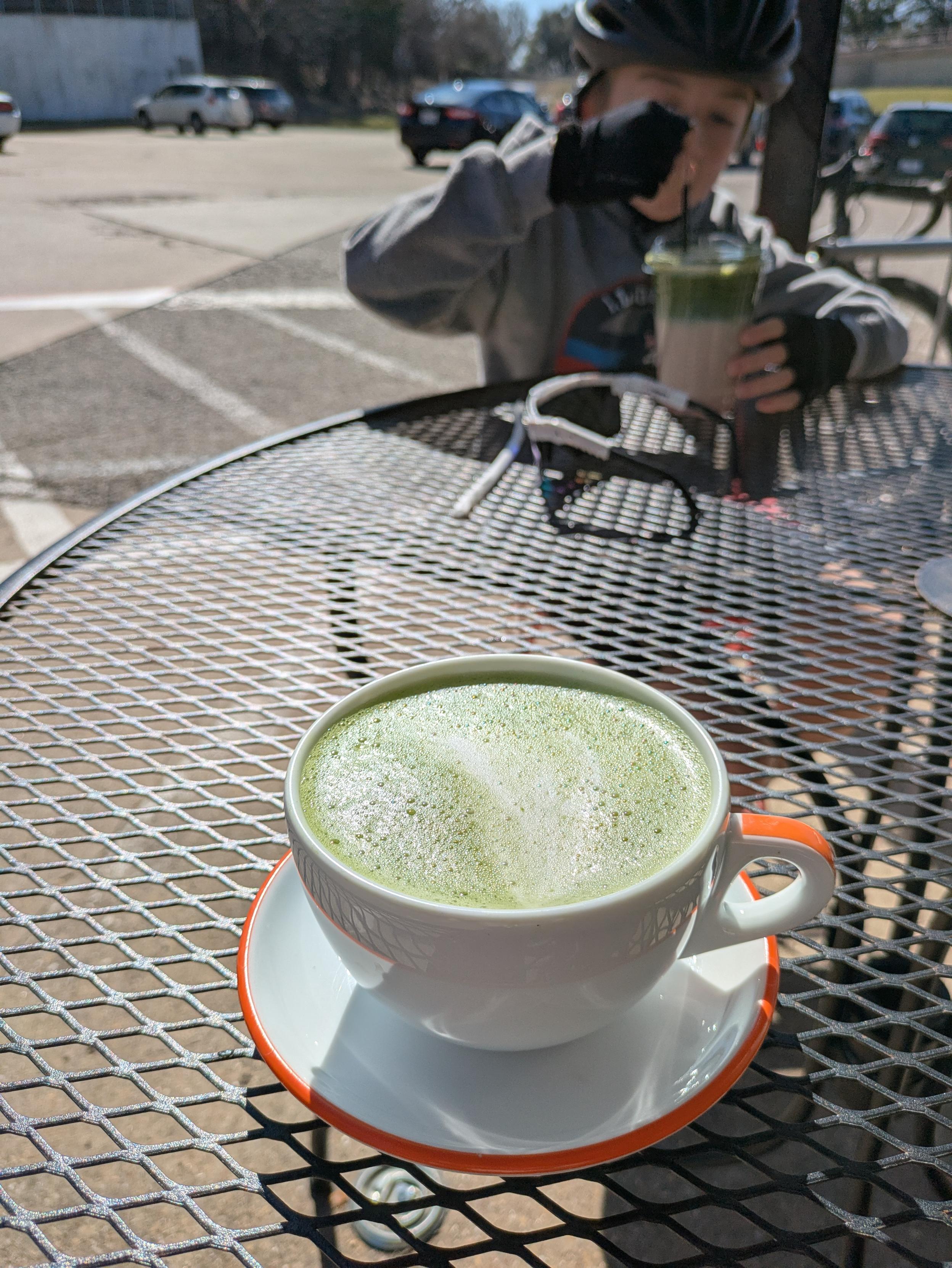 Foreground: a matcha latte in a white mug with orange on the handle, in a saucer with orange around the edge 

Background: a boy wearing a bike helmet stirring an iced matcha drink 