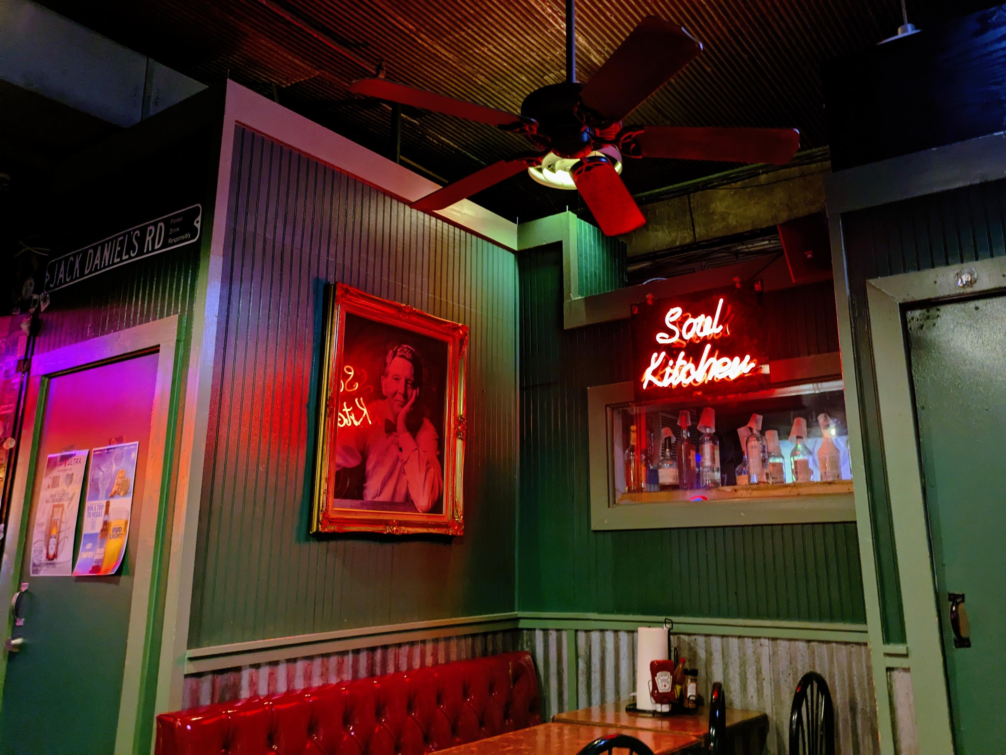 The image showcases the interior of a dimly lit bar or restaurant with a retro, Southern-inspired ambiance. The walls are painted dark green with vertical paneling, and there are red and purple lighting accents. A red neon sign reading "Soul Kitchen" is mounted above a small window that displays liquor bottles arranged in a row, some with cups placed on top.

A framed portrait of a smiling man in a white shirt hangs on the wall, reflecting the neon sign’s red glow. To the left of the portrait, a street-style sign reads "JACK DANIEL'S RD," adding to the whiskey bar aesthetic. Below the portrait is a red tufted leather bench along the wall, accompanied by a wooden table and black chairs.

The ceiling features a wooden corrugated texture, with a ceiling fan that has dark red blades hanging from it. The overall decor, with rustic materials, neon lighting, and vintage-style wall adornments, gives the space a nostalgic and inviting feel, reminiscent of a classic Southern bar or diner.