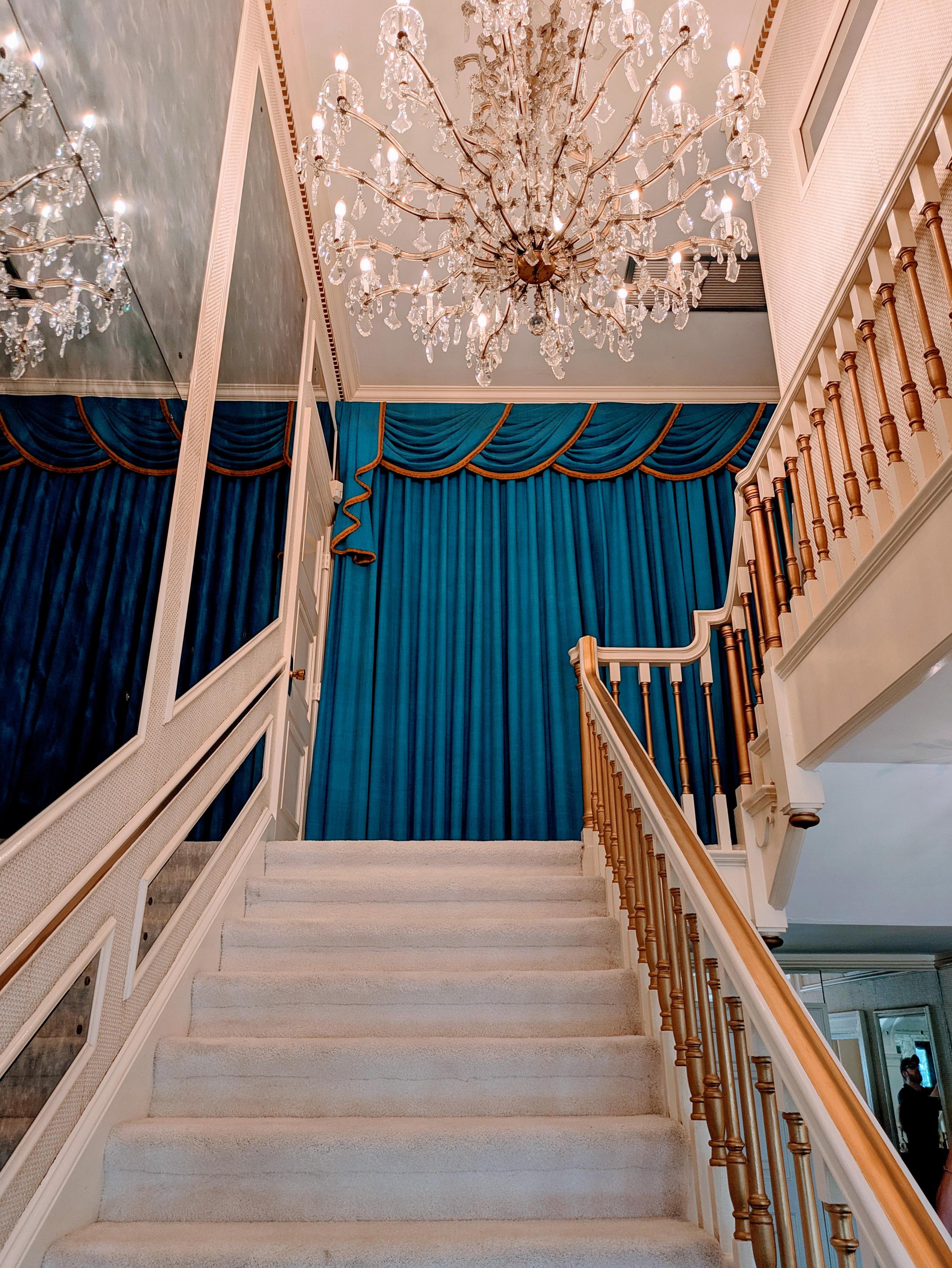 Looking up the stairs at Elvis' house at Graceland. White carpet, gold banister, big chandelier, blue drapes across the wall at the top