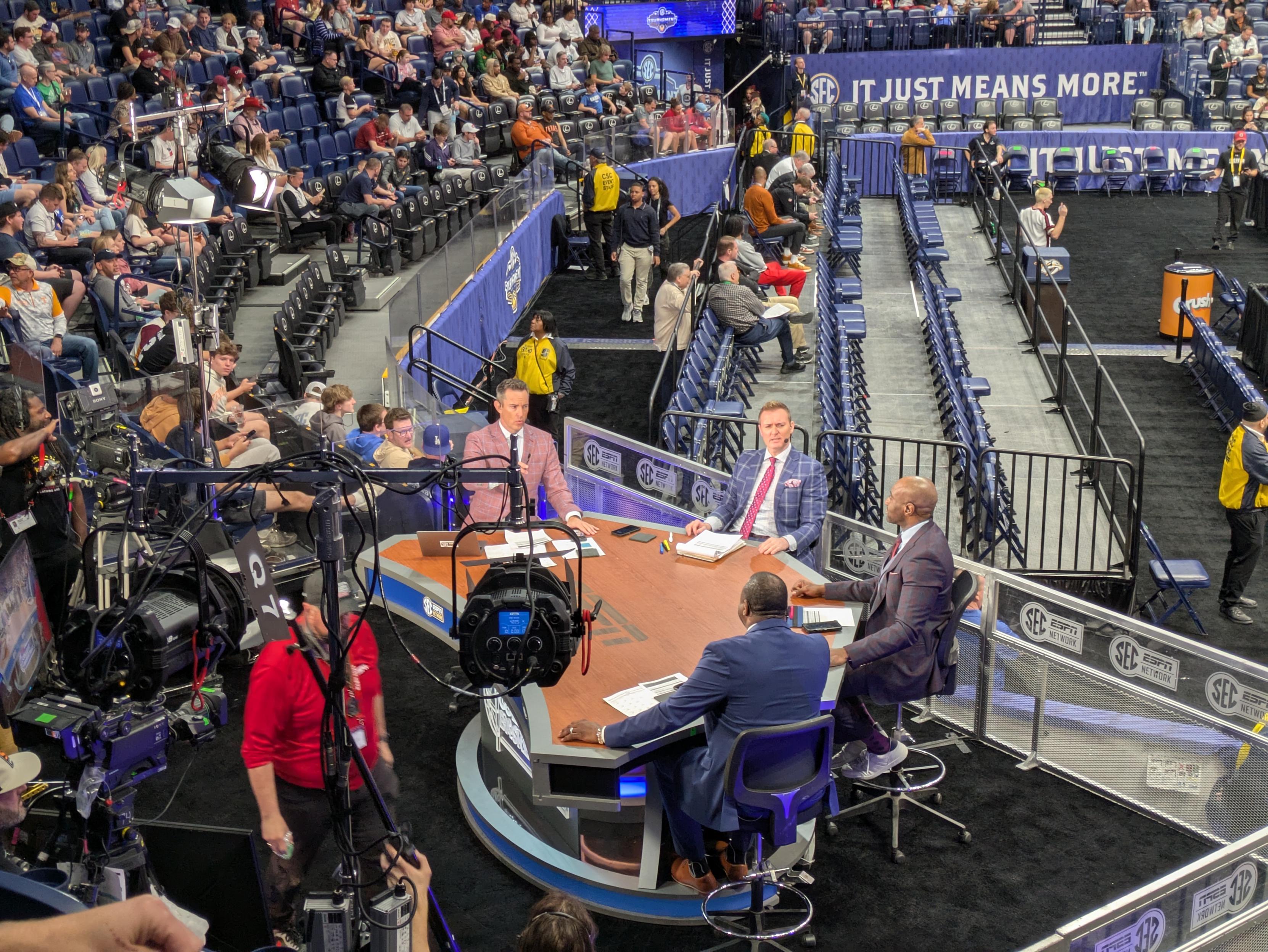 A photo of the ESPN broadcast desk in an arena, taken from above. Four basketball analysts sit around the desk, brightly lit and all wearing plaid suit jackets