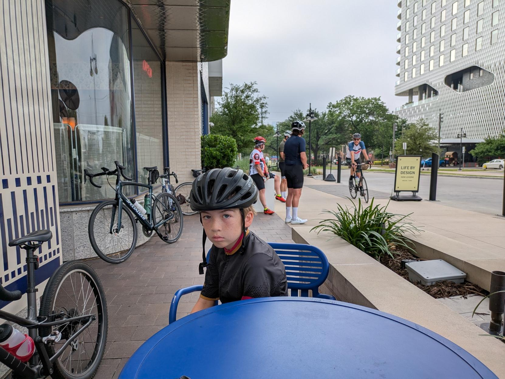 Boy sitting at cafe in cycling kit and helmet looking serious at the camera