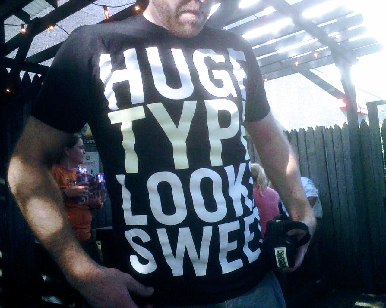 Photo of the author of this account from 2008, taken on a Blackberry phone, I'm wearing a shirt that says, "Huge type looks sweet," in large, all-caps letters.