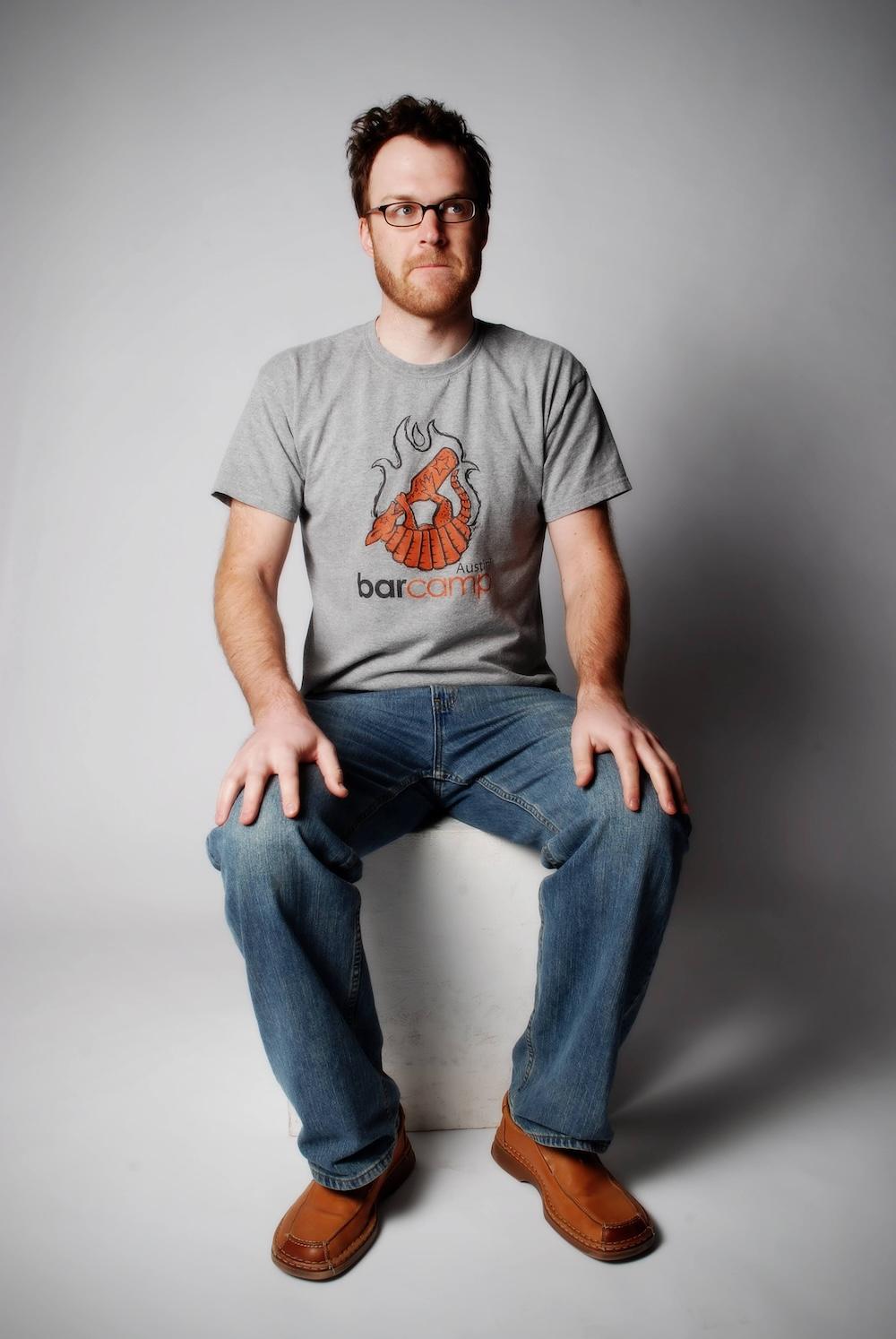 Photo of the owner of this account from approx 2008. He's sitting on a white block, wearing a grey shirt that says "barcamp Austin" depicting an armadillo drinking from a bottle