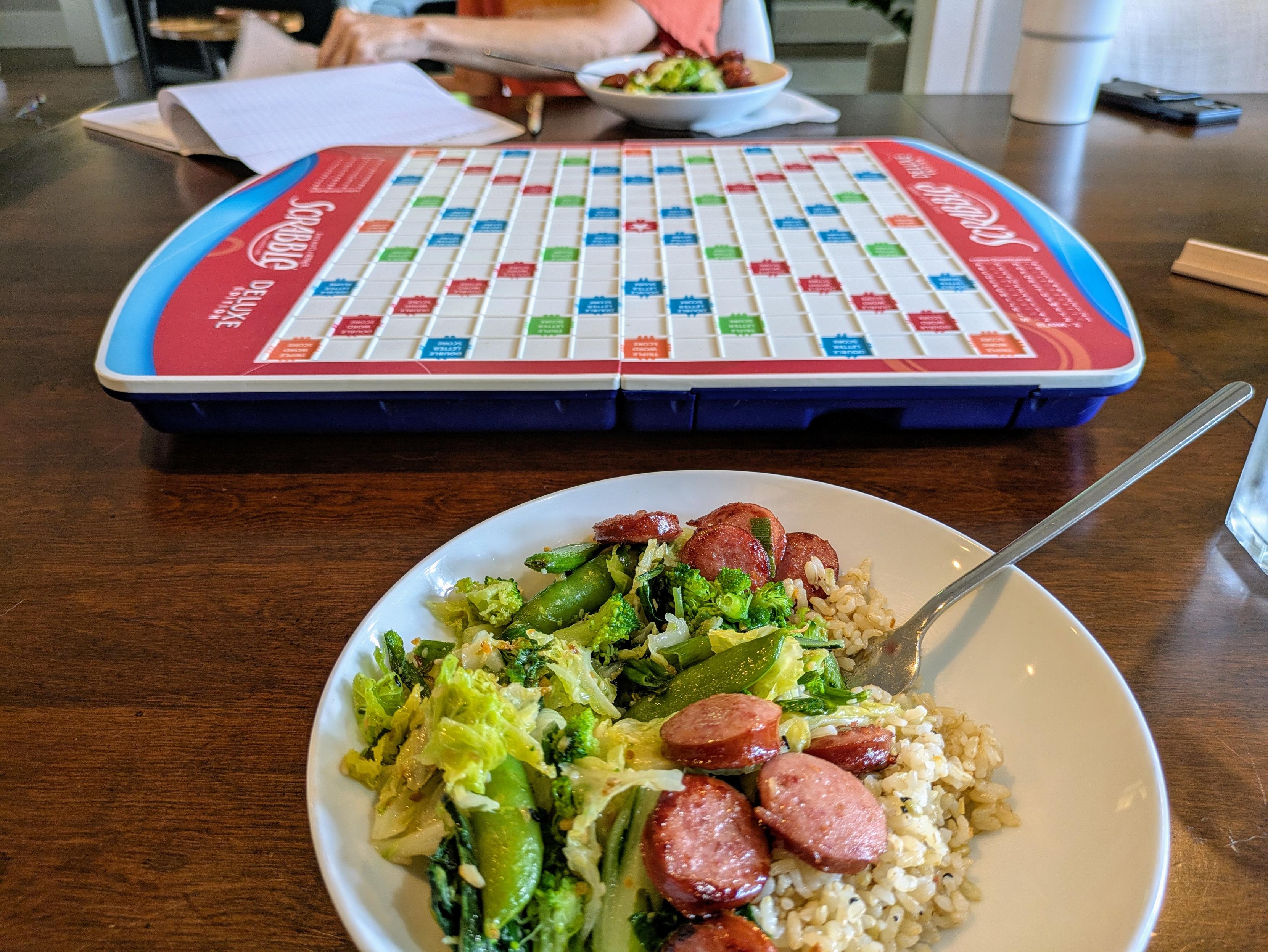 Foreground: A white bowl filled with a meal consisting of brown rice, sautéed sausage slices, and a mix of green vegetables like broccoli, cabbage, and snap peas. A fork is placed in the bowl.

Center: A Scrabble Deluxe Edition game board, currently empty, sits between two players on a wooden table.

Background: Another person (partially visible, wearing an orange shirt) is seated on the other side of the table, also with a similar bowl of food. There's a notebook or paper in front of them, possibly being reviewed or written on.