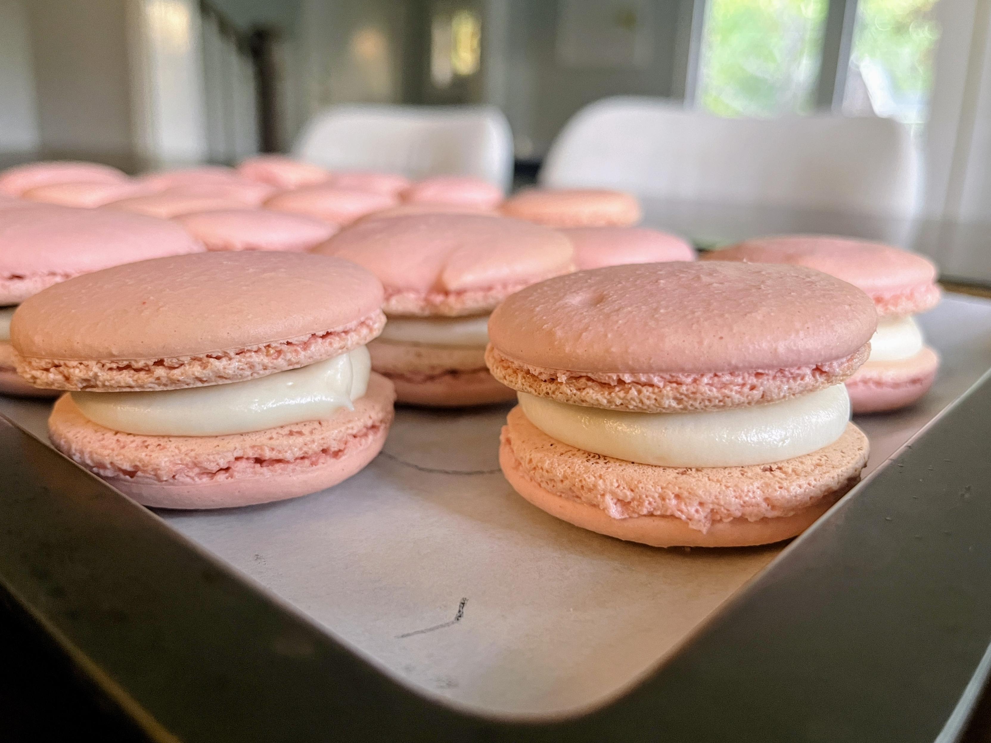 Pink macarons on a baking tray lined with parchment paper. Each has two smooth shells with ruffled edges and a white cream filling. The photo is taken indoors with natural light from a window in the background.