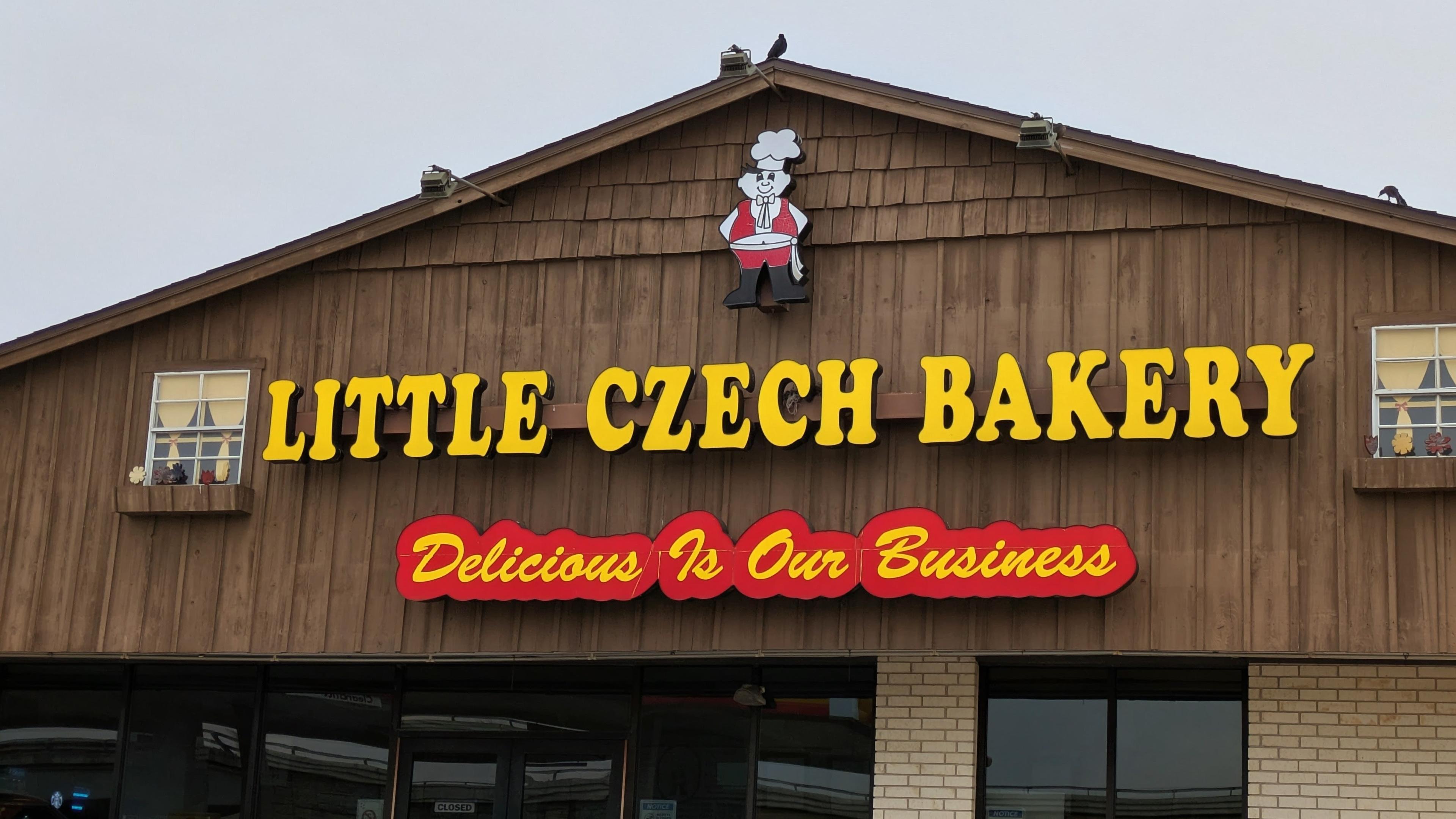 The image shows the front of Little Czech Bakery, a building with a wood exterior and brick lower wall. The sign reads “LITTLE CZECH BAKERY” in yellow block letters and “Delicious Is Our Business” in red script. Above the sign is a cartoon baker wearing a chef’s hat and red outfit.