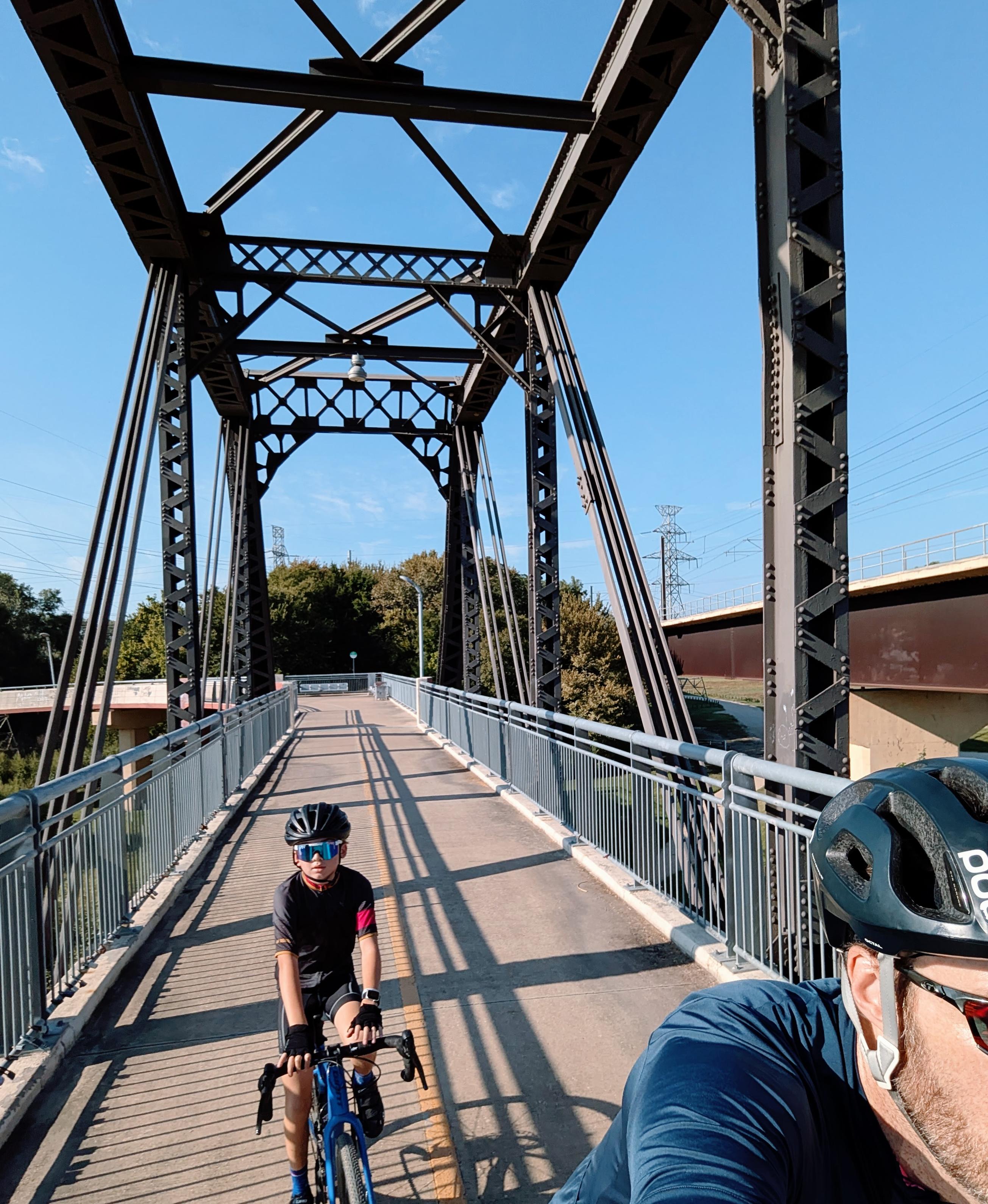 A child on a bike follows an adult across a steel truss bridge on a sunny day.