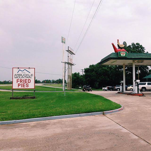 A Sinclair gas station with the red dinosaur and a chicken perched above the pumps, and a sign that says Arbuckle Mountain Fried Pies