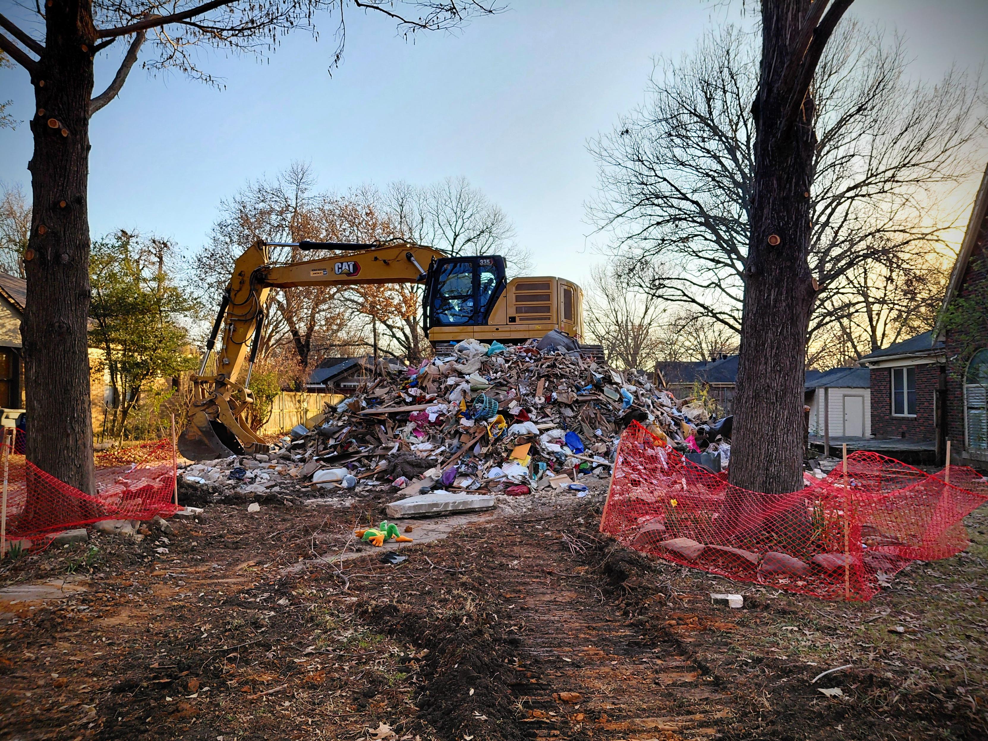 A yellow excavator sits atop a large pile of mixed demolition debris—wood, insulation, plastic, and household items—in a residential lot at sunset. Bare winter trees frame the scene, and red plastic construction fencing surrounds parts of the cleared ground. Tire tracks cut through the dirt in the foreground, with neighboring brick houses visible in the background.